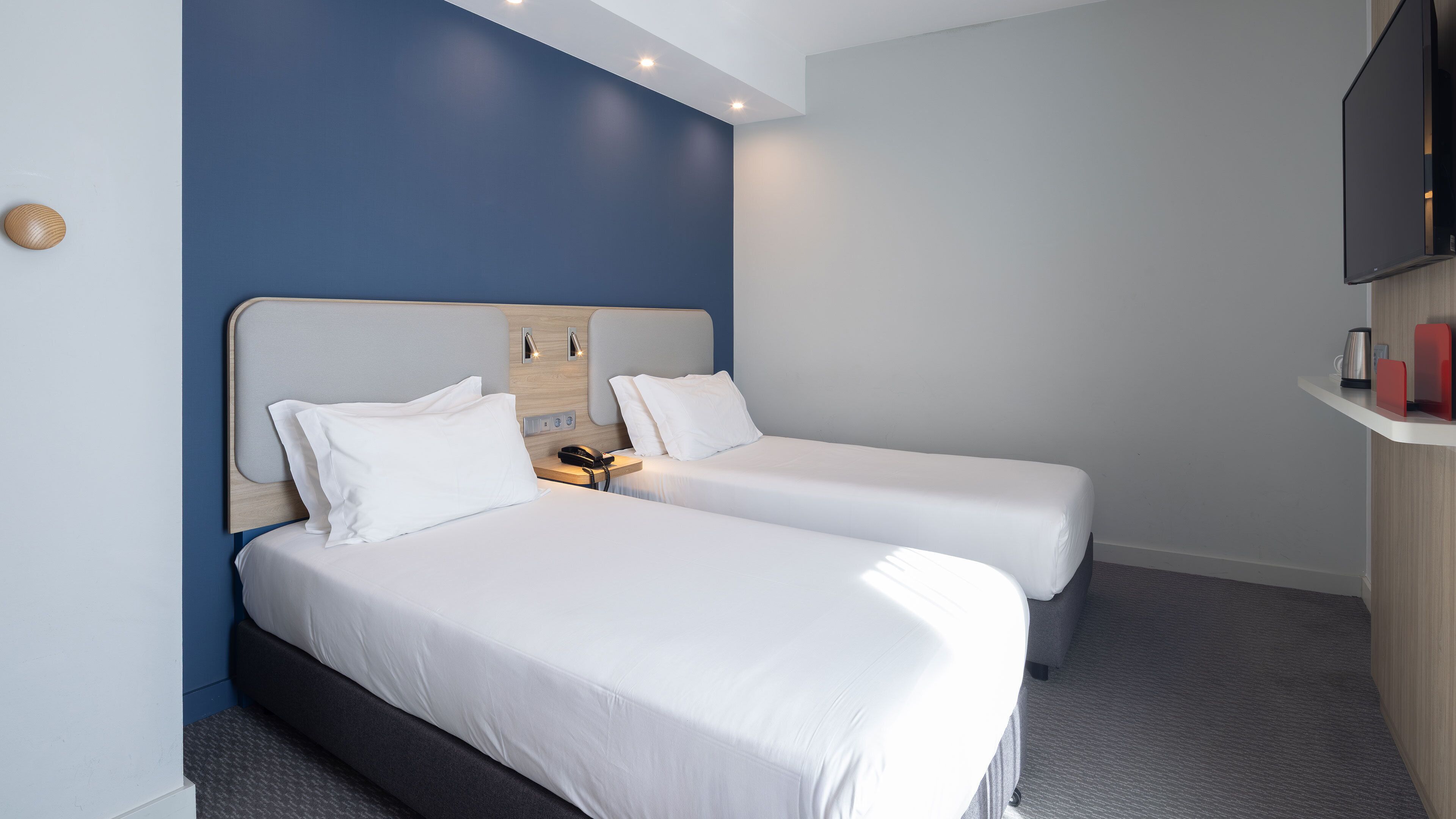 A hotel room with two beds, a wooden headboard, a flat-screen TV, and a small shelf with a kettle and a red object.