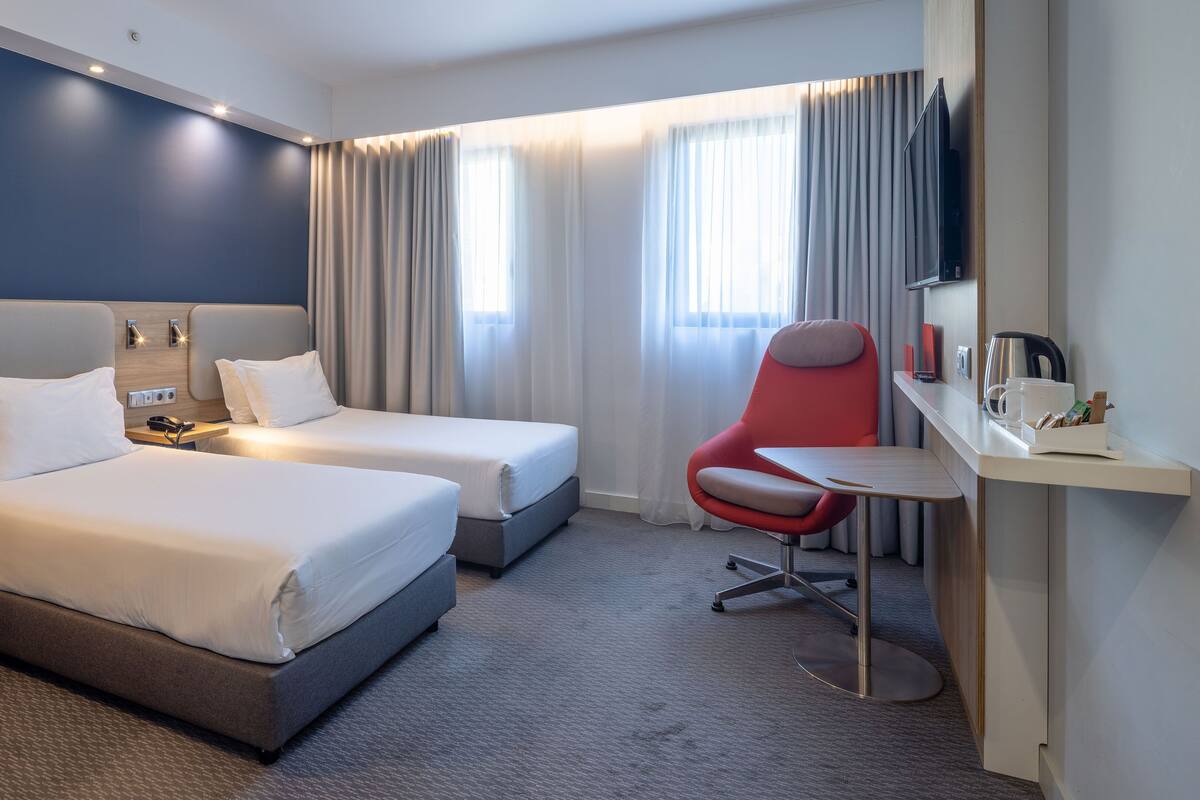 Hotellihuone, jossa on kaksi sänkyä, työpöytä tuolilla, televisio ja ikkuna, jossa on verhot.