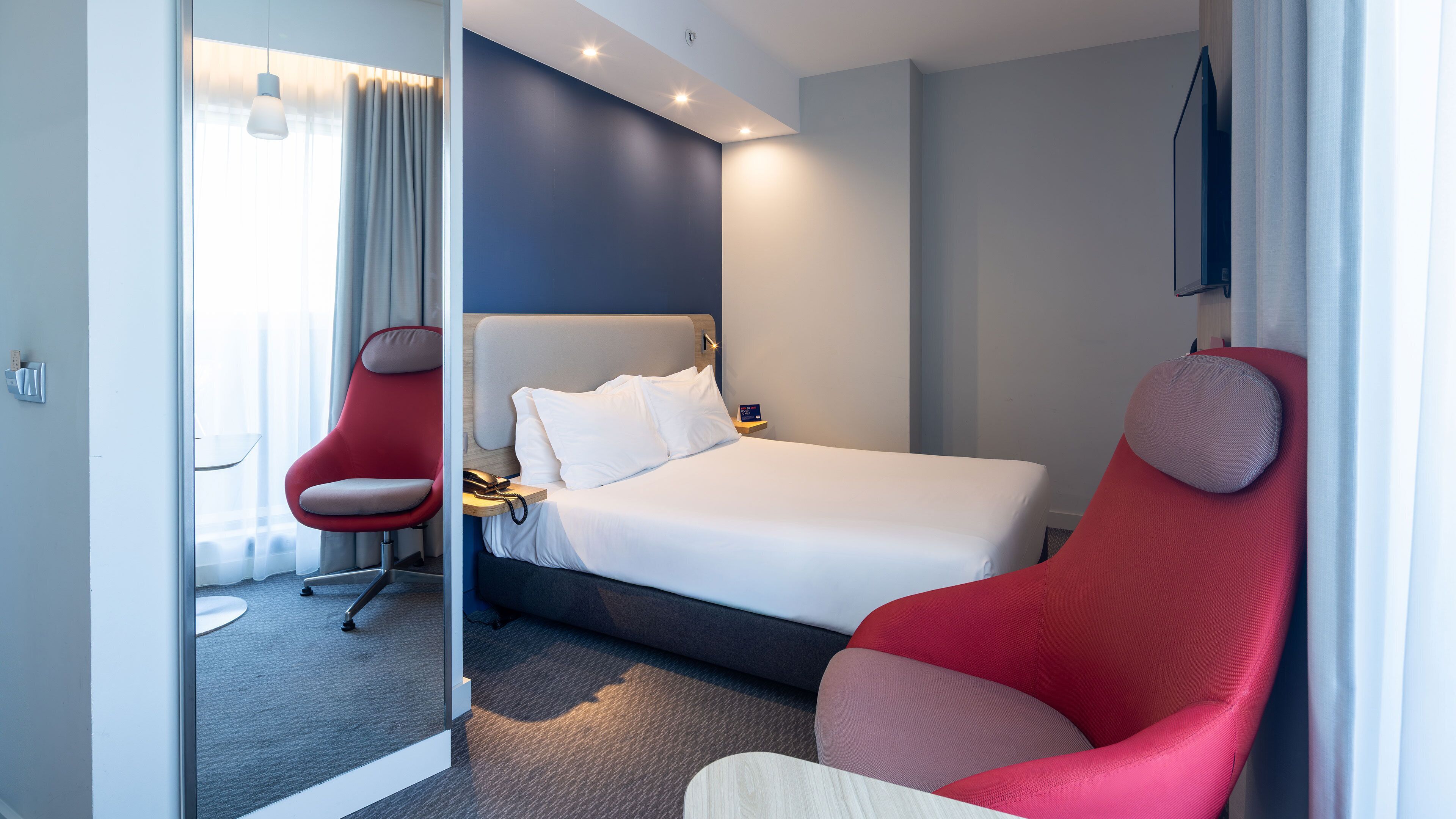 Et moderne hotelværelse med en seng, en rød lænestol, et skrivebord med telefon og et spejl.