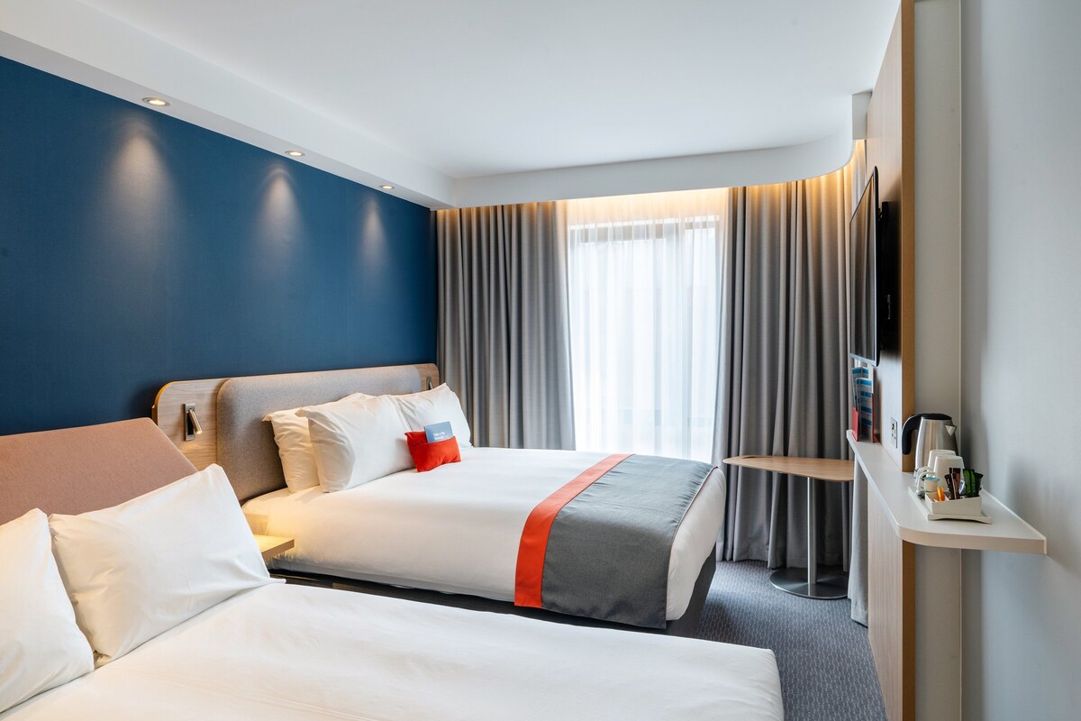 A hotel room with two beds, a desk, a TV, and a window with curtains.