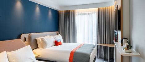 Et hotelværelse med to senge, et skrivebord, et fjernsyn og et vindue med gardiner.