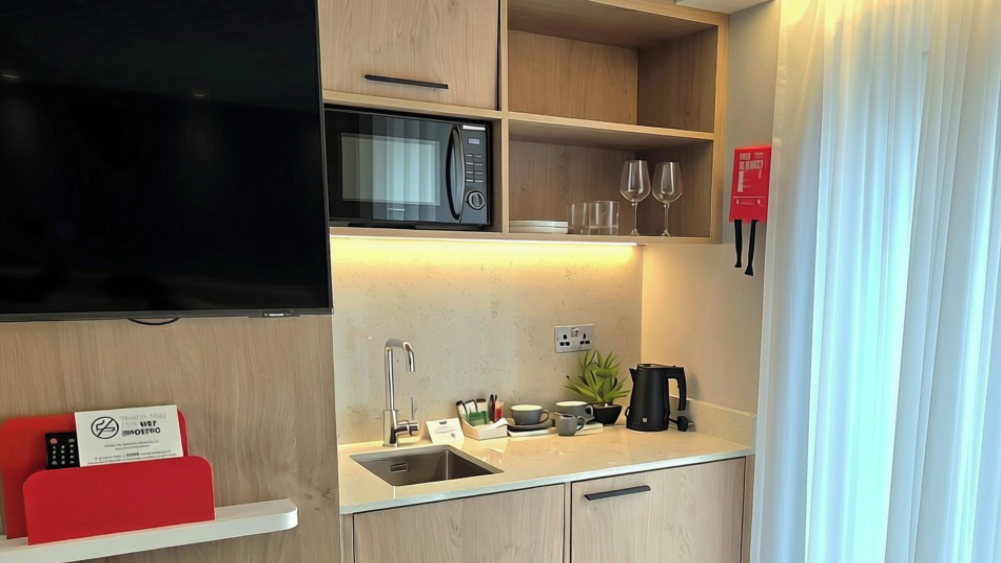 A compact kitchenette with a microwave, a sink, and a coffee maker. There is a small shelf with a red object and a sign, and a television mounted on the wall.