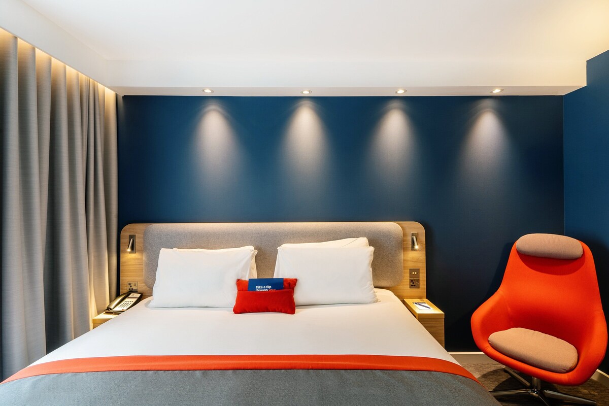A modern hotel room with a large bed, a red chair, and a bedside table with a phone and a book.