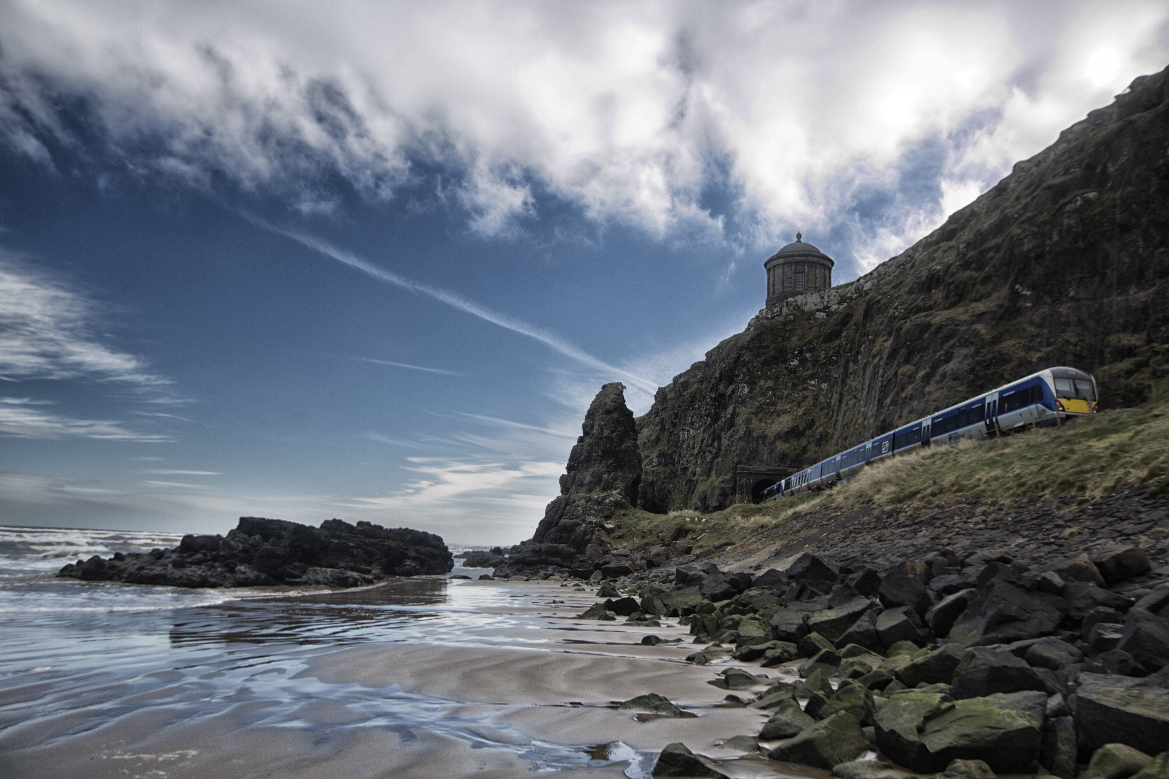 A coastal landscape with a train on a rocky embankment, a clear sky, and a body of water.