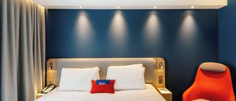 Una camera d'albergo moderna con un letto grande, una poltrona rossa e un comodino con un telefono e un libro.