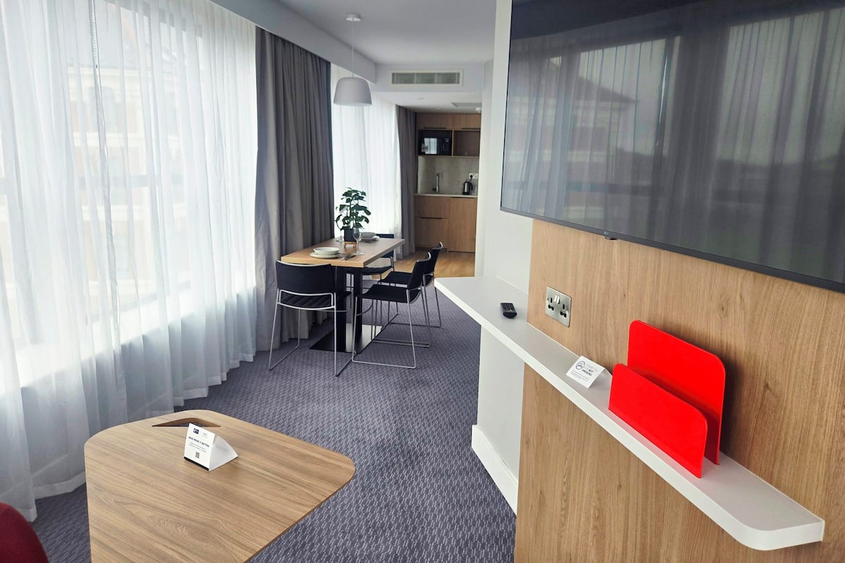 Habitación de hotel moderna con escritorio de madera, pared de acento roja, televisor montado y zona de comedor con planta y cortinas.