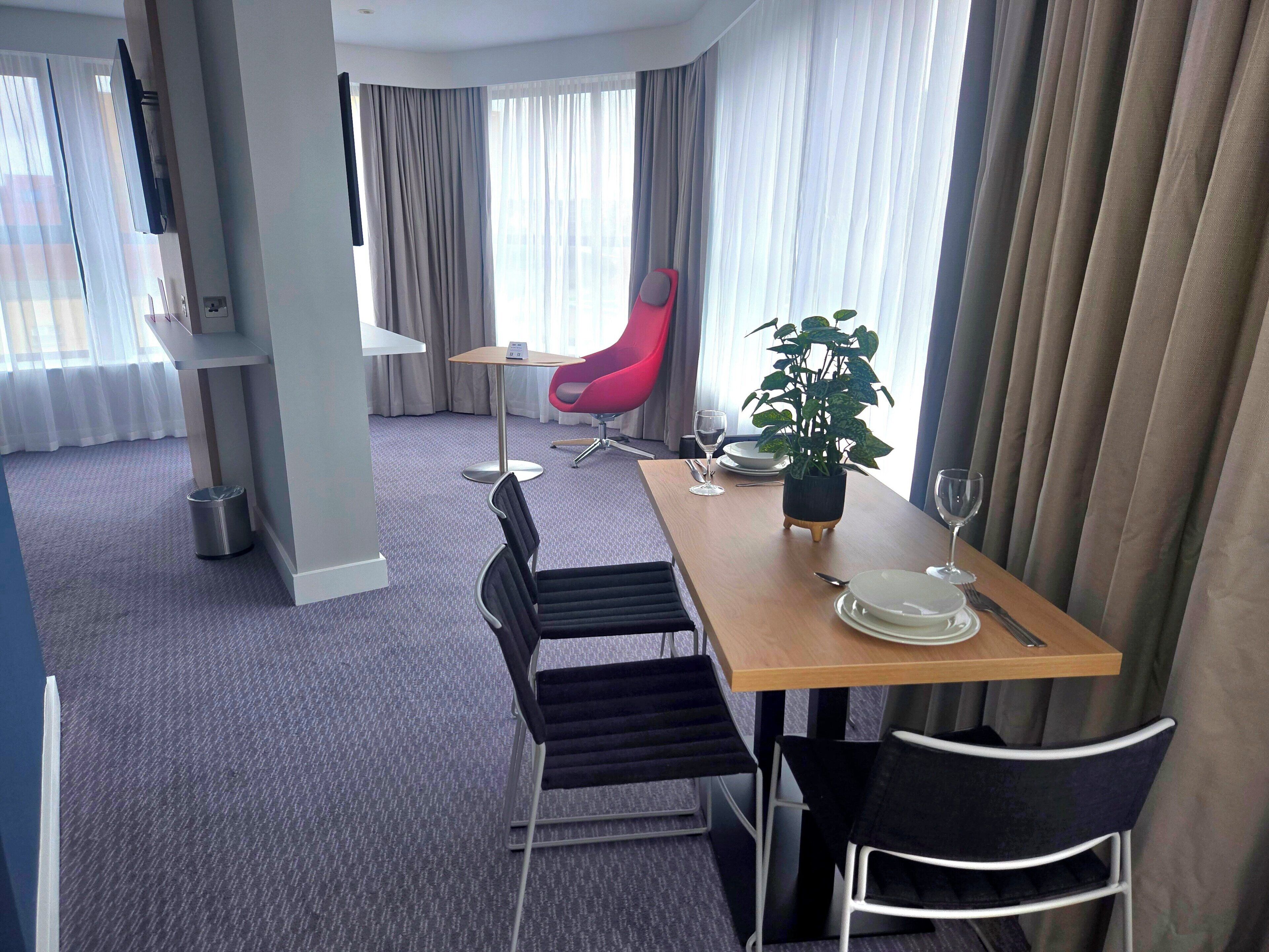A hotel room with a dining area featuring a wooden table, black chairs, a red chair, a potted plant, and a TV mounted on the wall.
