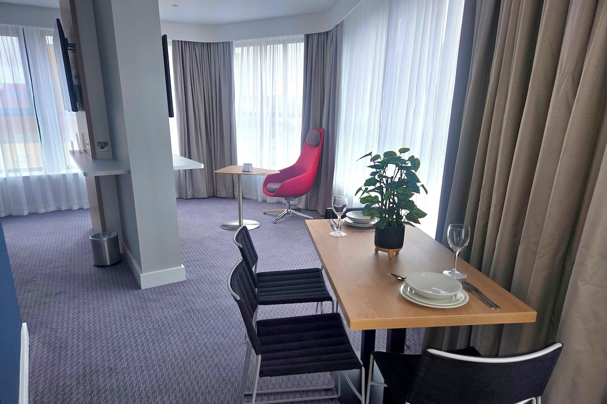A hotel room with a dining area featuring a wooden table, black chairs, a red chair, a potted plant, and a TV mounted on the wall.