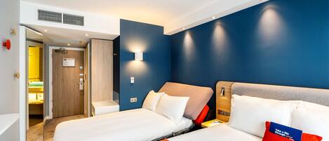 A hotel room with two beds, a blue accent wall, a wooden headboard, and a red pillow with text.