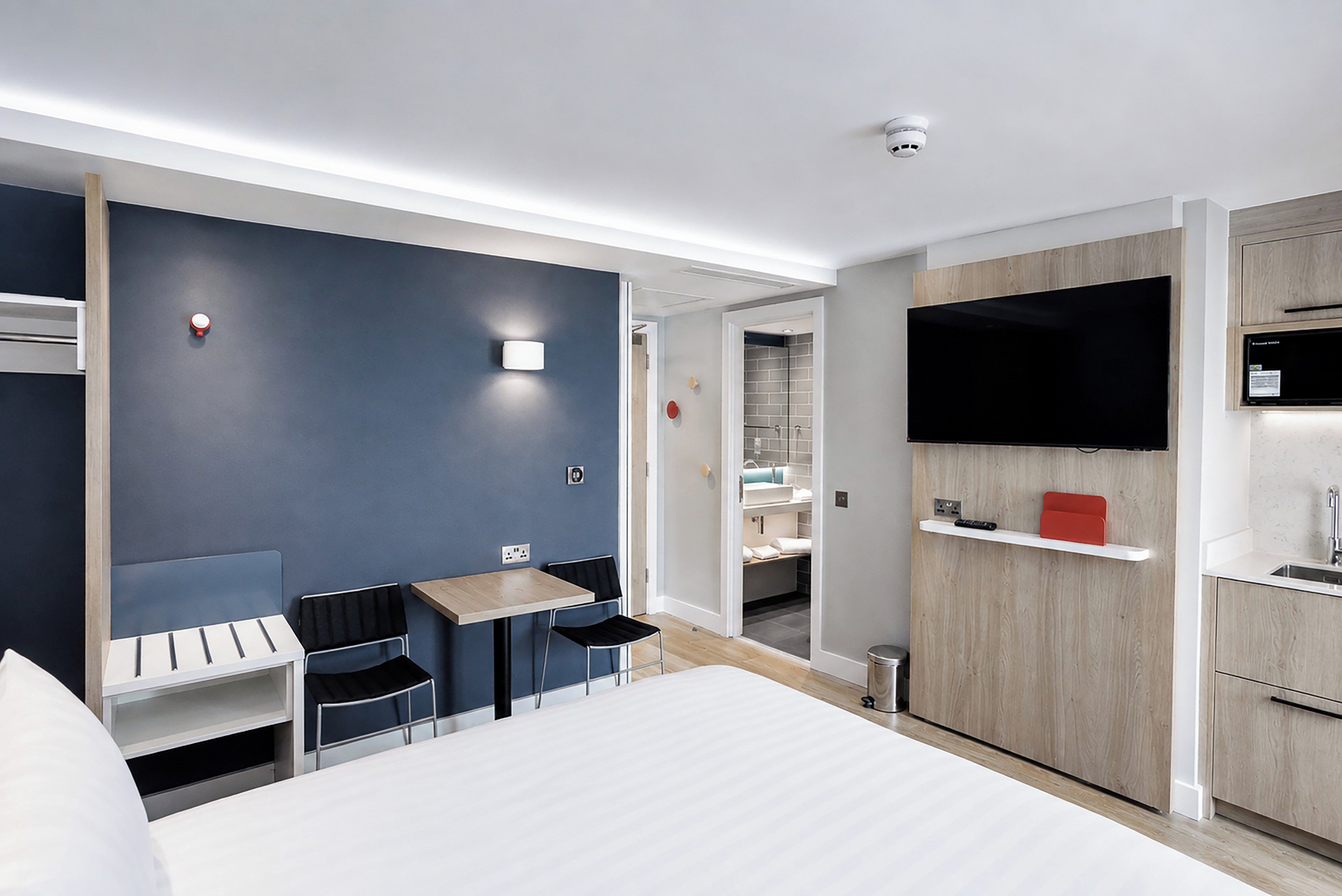 A modern hotel room with a bed, a desk, two chairs, a TV, and a kitchenette.