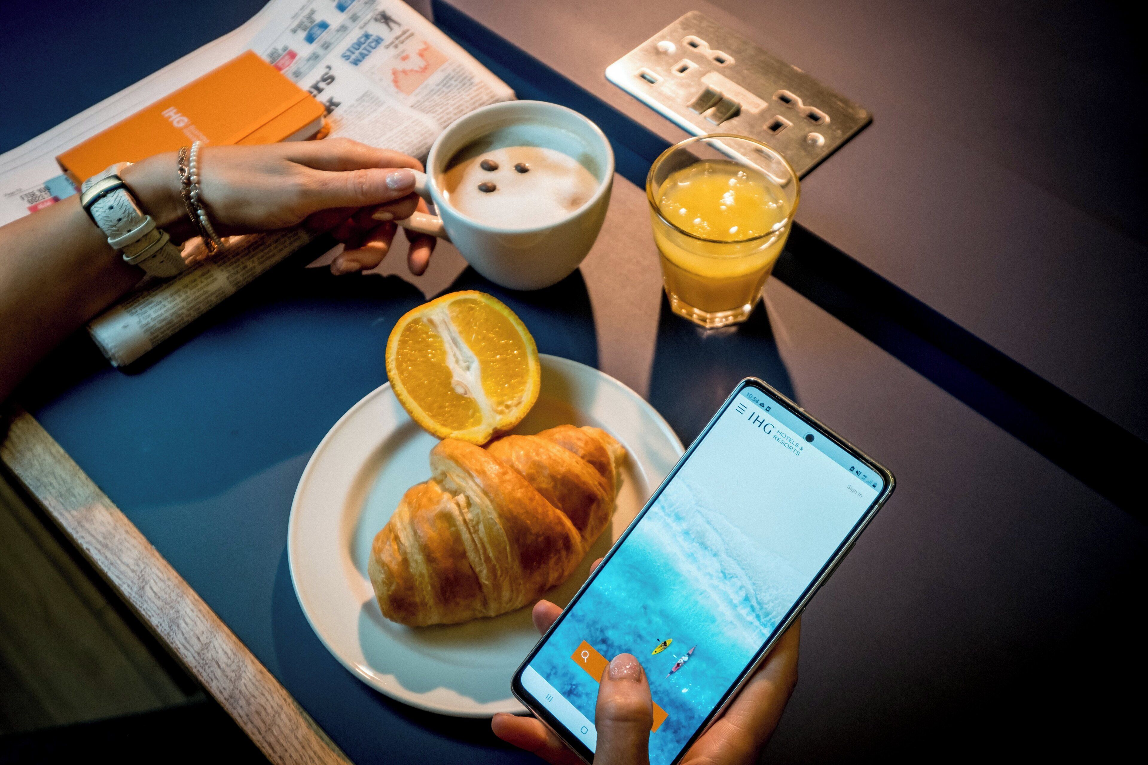 Una persona sosteniendo un teléfono inteligente con una aplicación de reservas de hotel, un croissant, un vaso de zumo de naranja y una taza de café sobre una mesa.