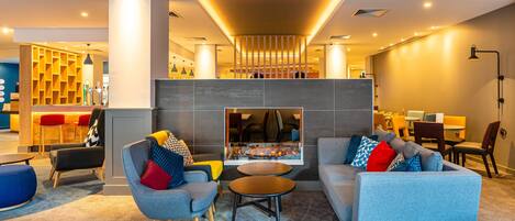 A modern lobby with a grey sofa, armchairs, and a small table. There is a bar area in the background and a seating area with a view of the lobby.