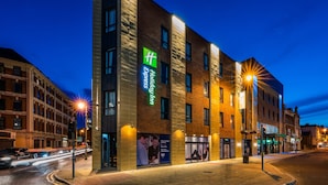 A modern hotel with a prominent green and white sign, large windows, and a well-lit facade.