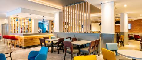 Un restaurante moderno con una zona de bar, múltiples opciones de asientos, incluyendo mesas redondas y sillas, y una cocina visible al fondo.