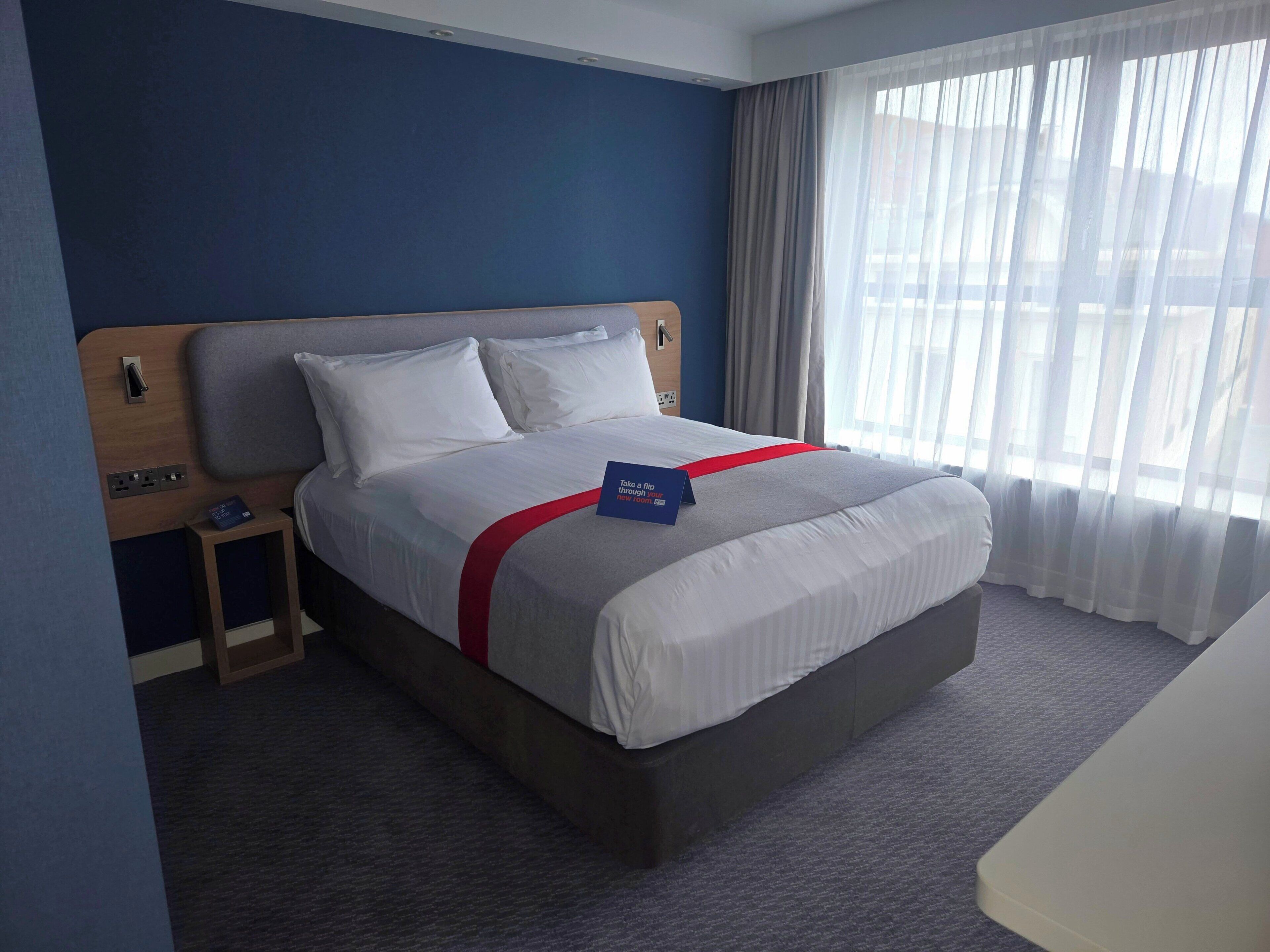 Una habitación de hotel con una cama, una mesita de noche, una colcha roja y blanca, un cartel azul y una ventana con cortinas.