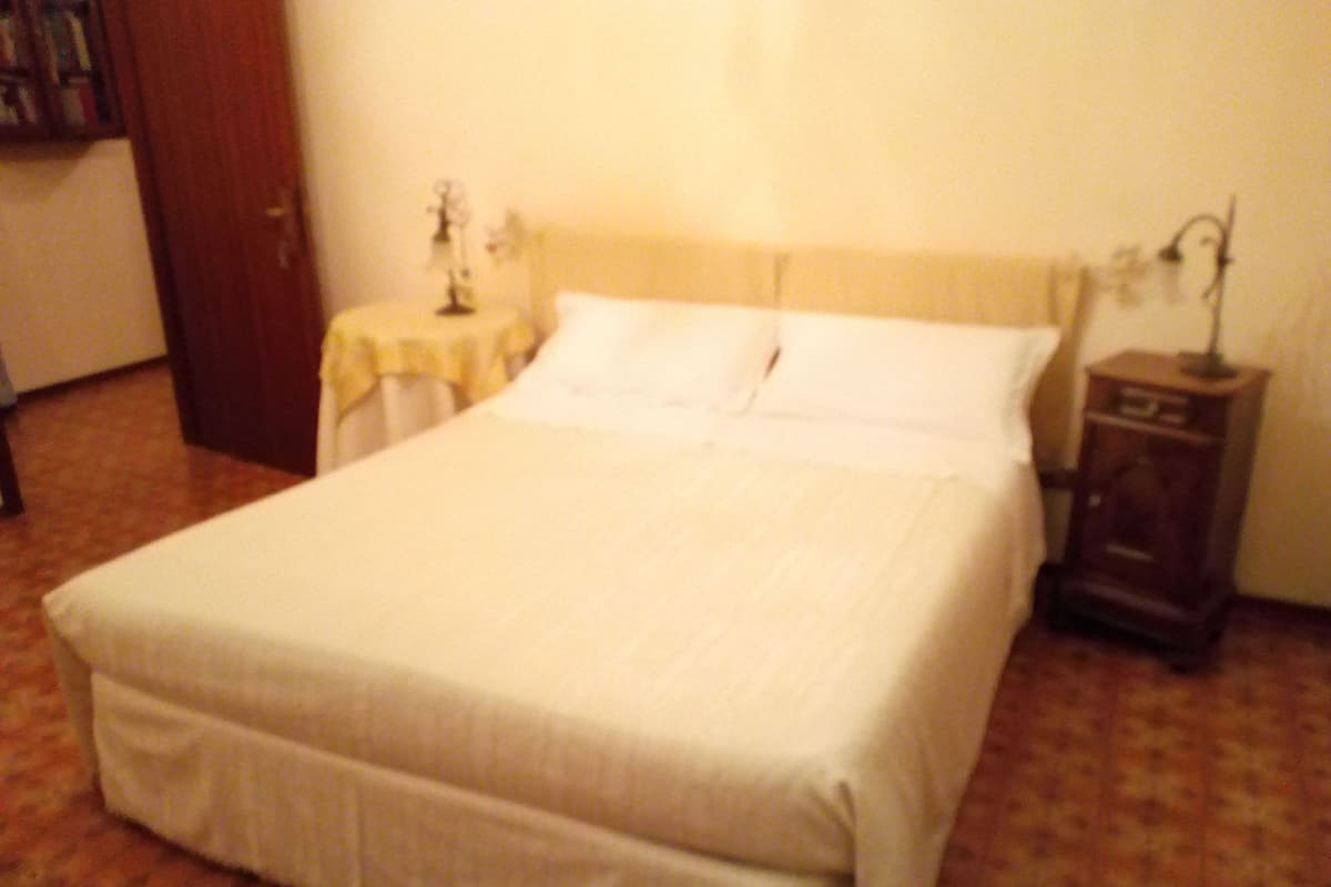 A neatly made bed with white linens in a room with a wooden floor and a small bedside table with a lamp.