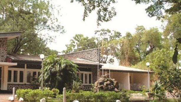 Un edificio con fachada de piedra, un acceso cubierto y un jardín con una estatua y plantas.