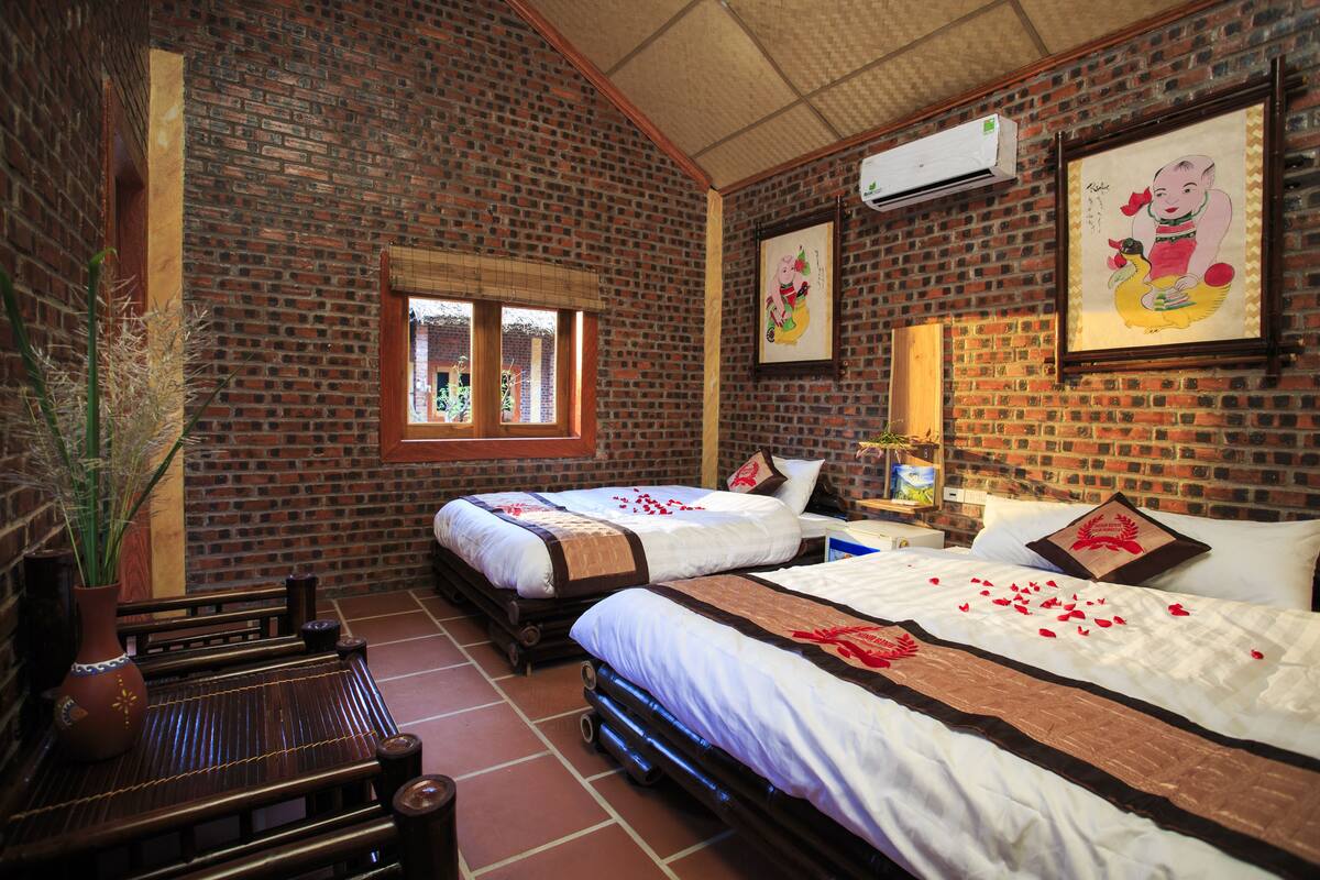 Een hotelkamer met bakstenen muren, twee bedden, een raam, een nachtkastje, een vaas en een wandairconditioner.