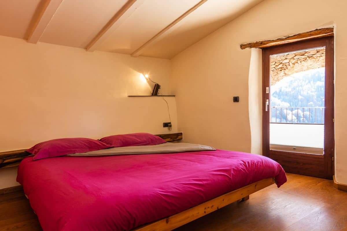 Un dormitorio con una cama de madera, un ventanal con vistas a las montañas y una estantería con una lámpara.