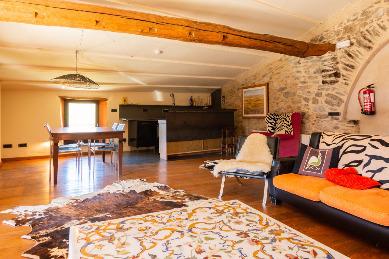 Un interior rústico con pared de piedra, vigas de madera, zona de comedor y un sofá con cojines estampados.
