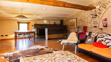 Un interior rústico con pared de piedra, vigas de madera, zona de comedor y un sofá con cojines estampados.