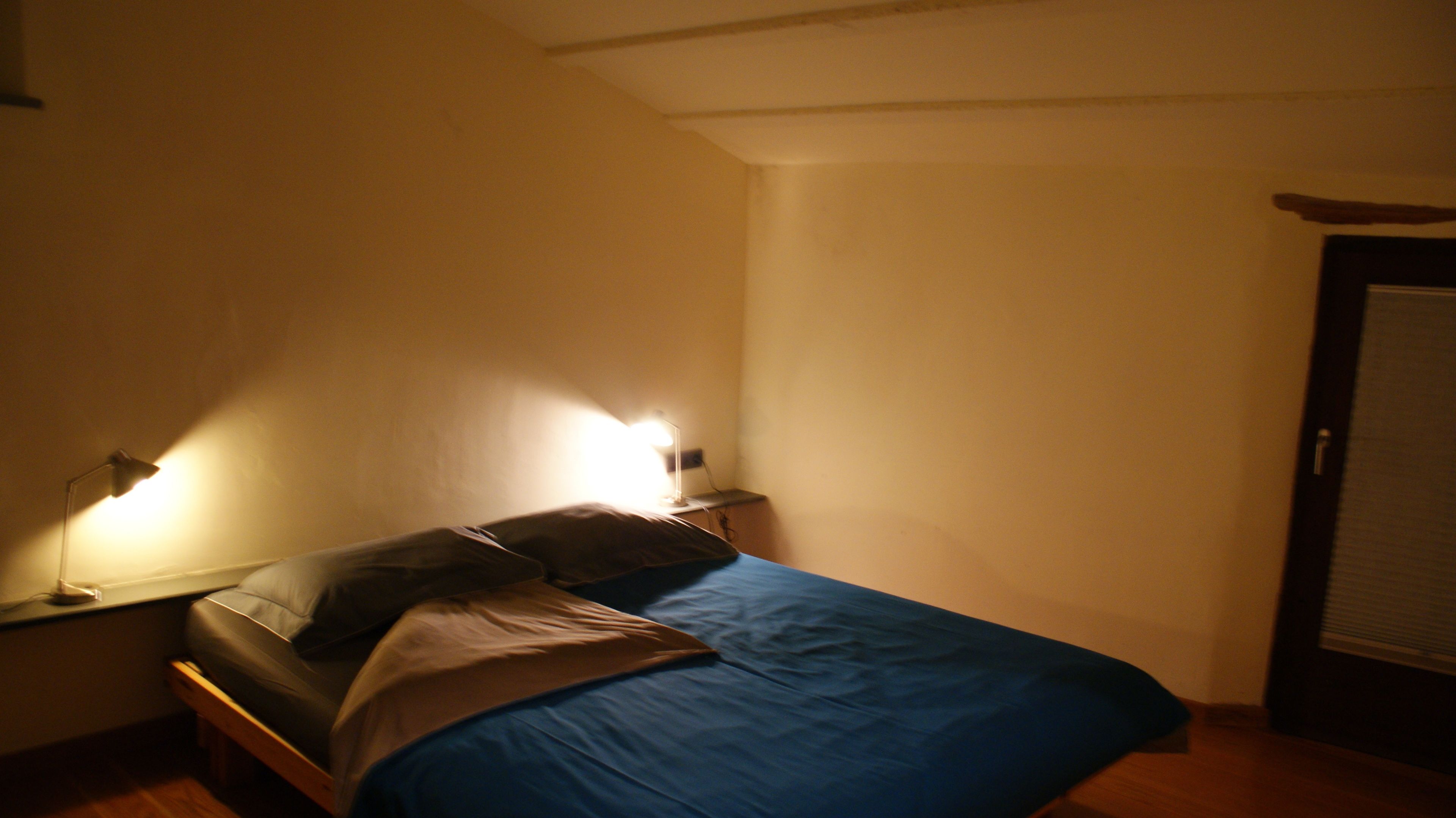 A small, dimly lit bedroom with a single bed, two bedside lamps, and a closed door.