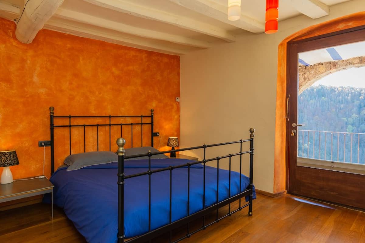 Un dormitorio con estructura de cama metálica, colcha azul, mesita de noche con lámpara y vistas a un paisaje montañoso a través de una puerta de madera.
