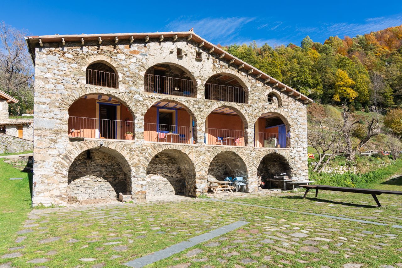 Un edificio de piedra con múltiples arcos y balcones, que cuenta con mesas al aire libre y una mesa de picnic.