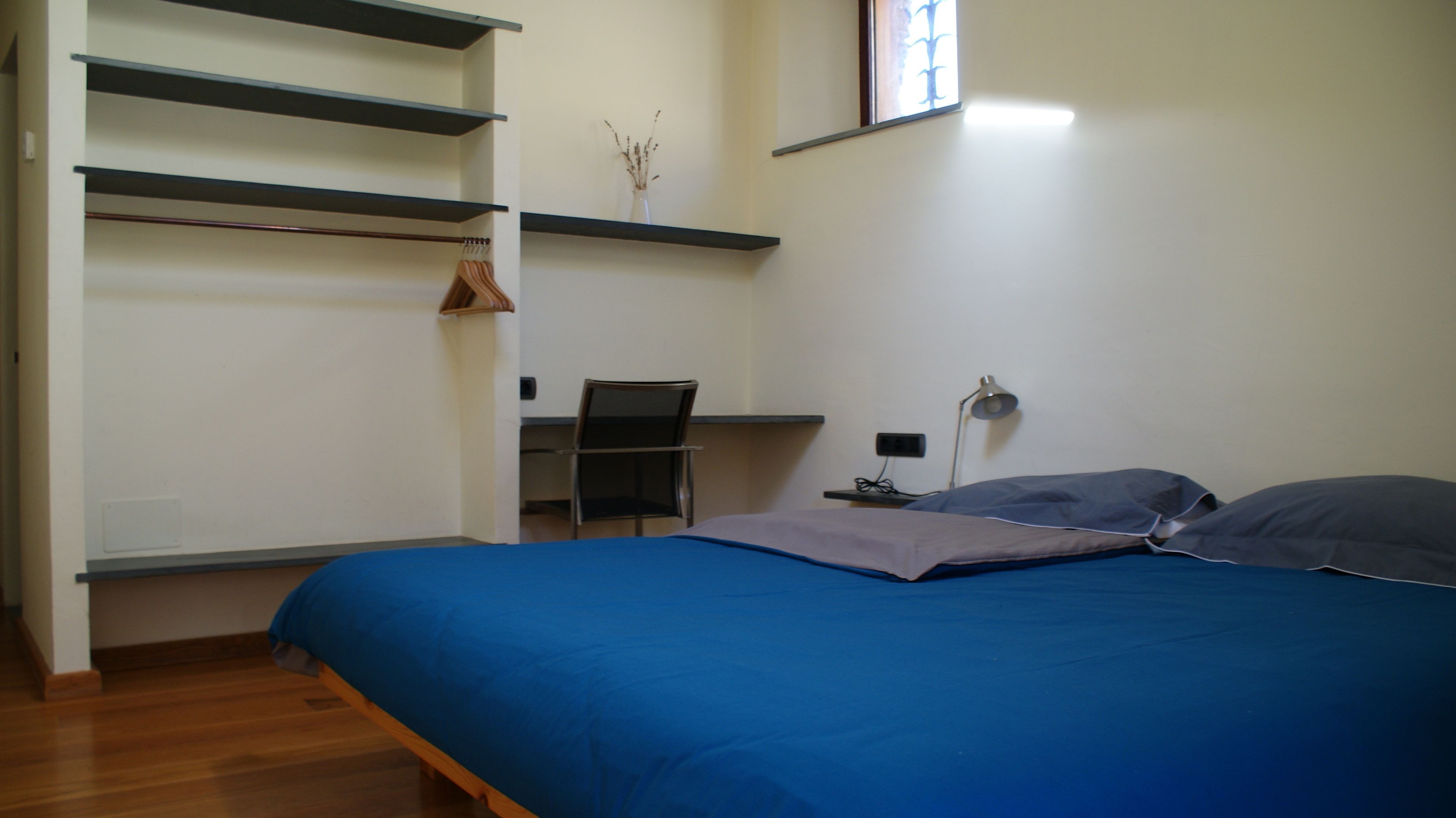 Une chambre à coucher avec un lit, un bureau et une chaise. Il y a une fenêtre avec des stores, une lampe et une étagère sur laquelle se trouve une plante.