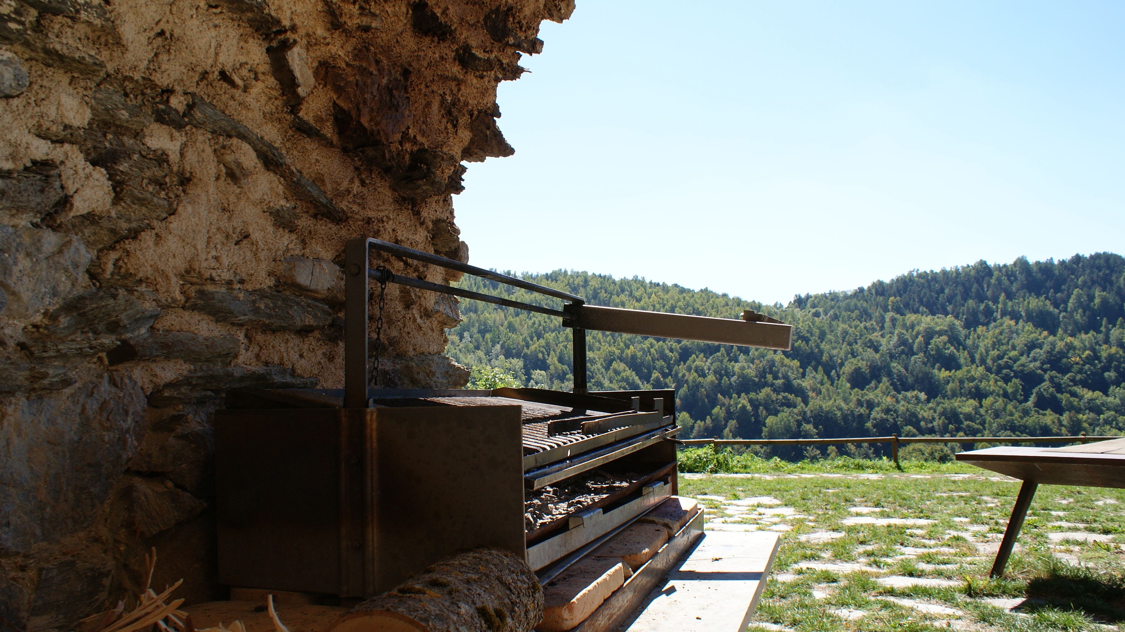 Una parrilla rústica al aire libre frente a un acantilado rocoso, con una montaña boscosa al fondo.