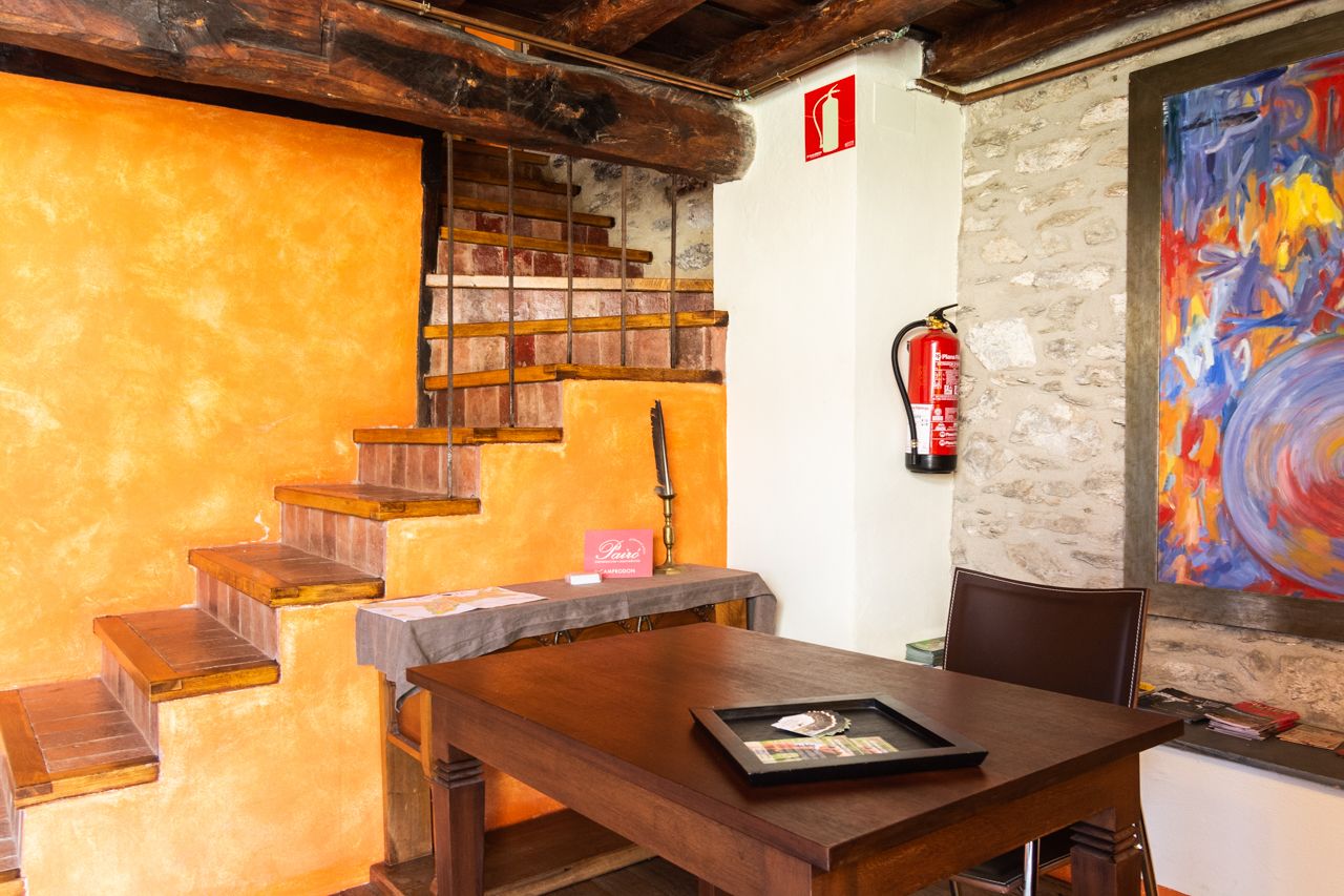 Una escalera de madera con un extintor montado en la pared, una mesa con una bandeja y un cuadro abstracto y colorido en la pared.