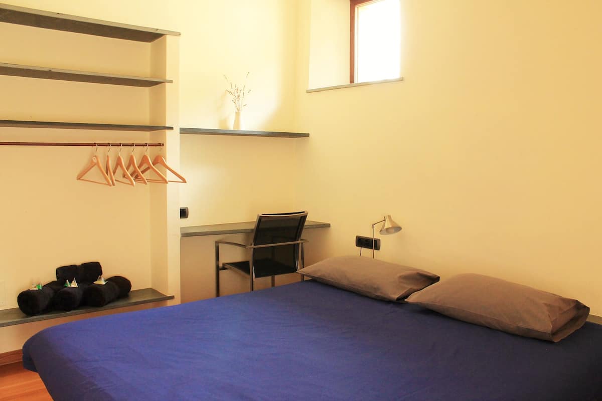Un dormitorio con cama, escritorio y silla. Hay perchas en un estante y una planta pequeña sobre el escritorio.