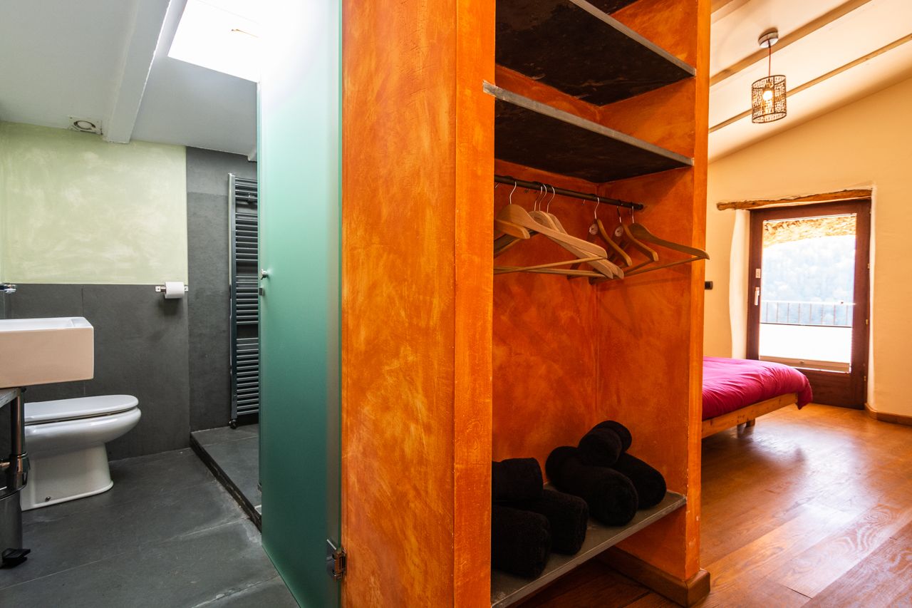 Habitación pequeña con armario de madera, cama, estante con zapatos, inodoro y ventana con vistas.