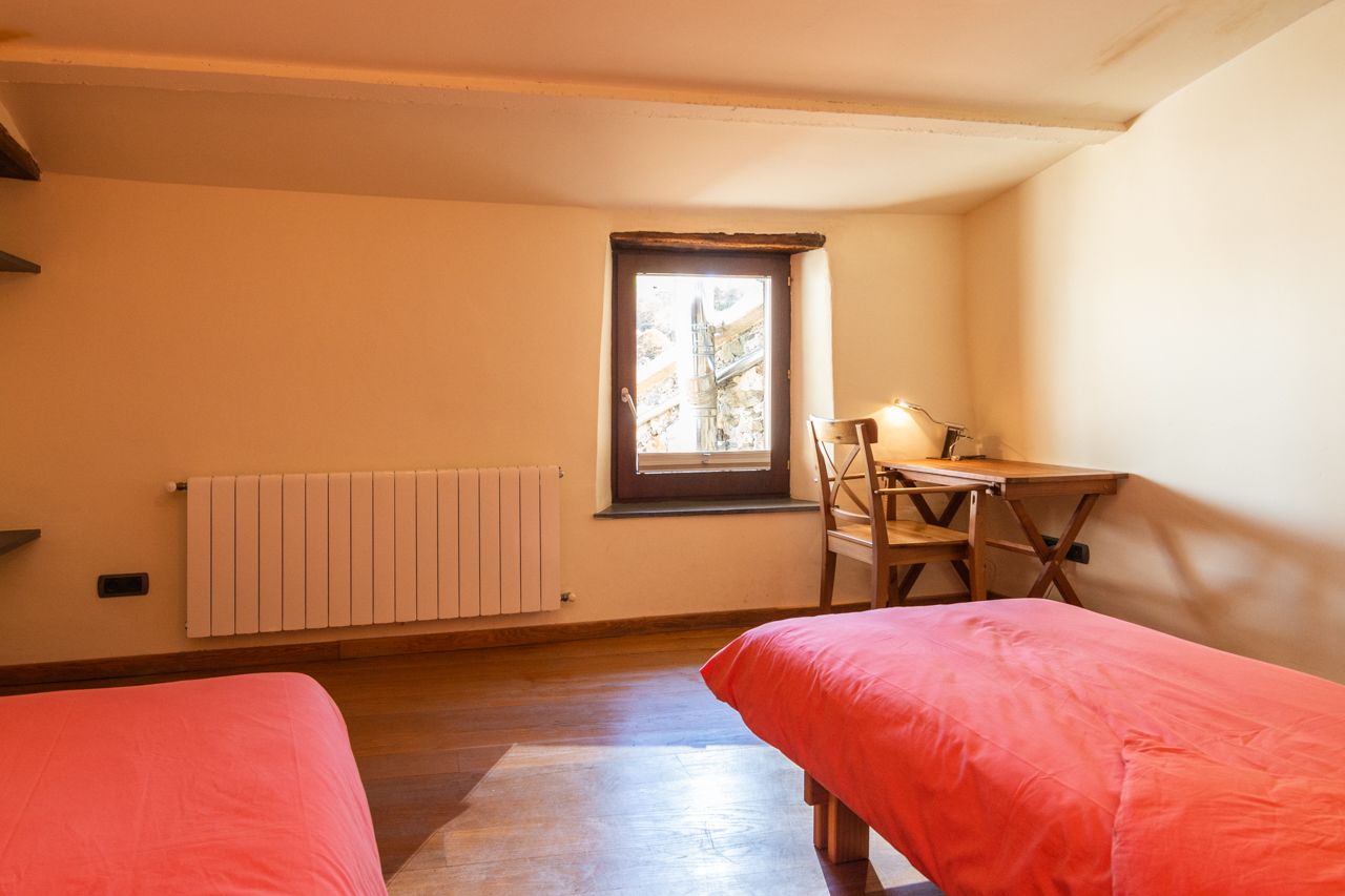 A bedroom with two beds, a desk with a chair, a radiator, and a window with a view of trees.