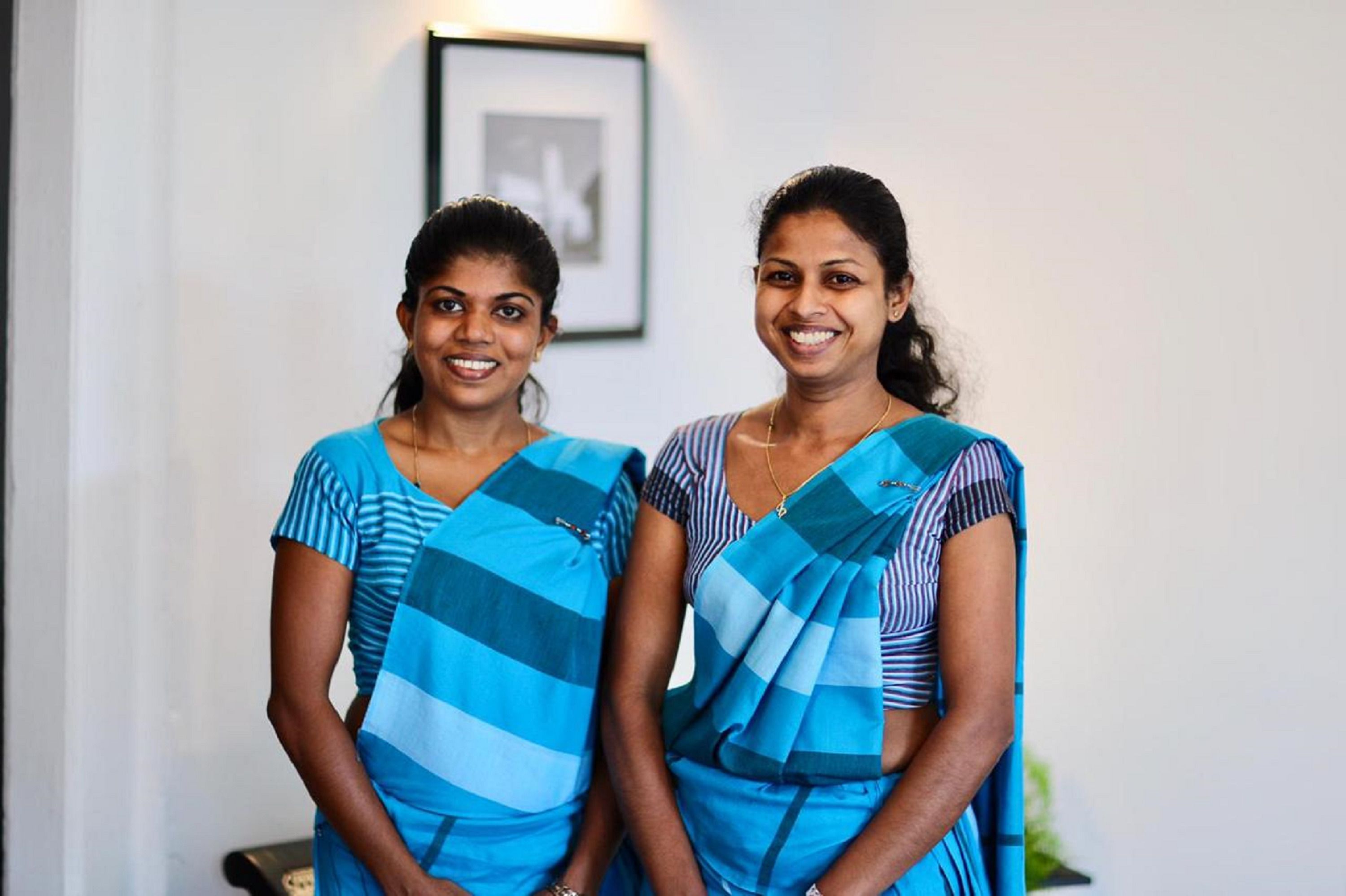 Two women in blue and white striped sarees standing indoors.