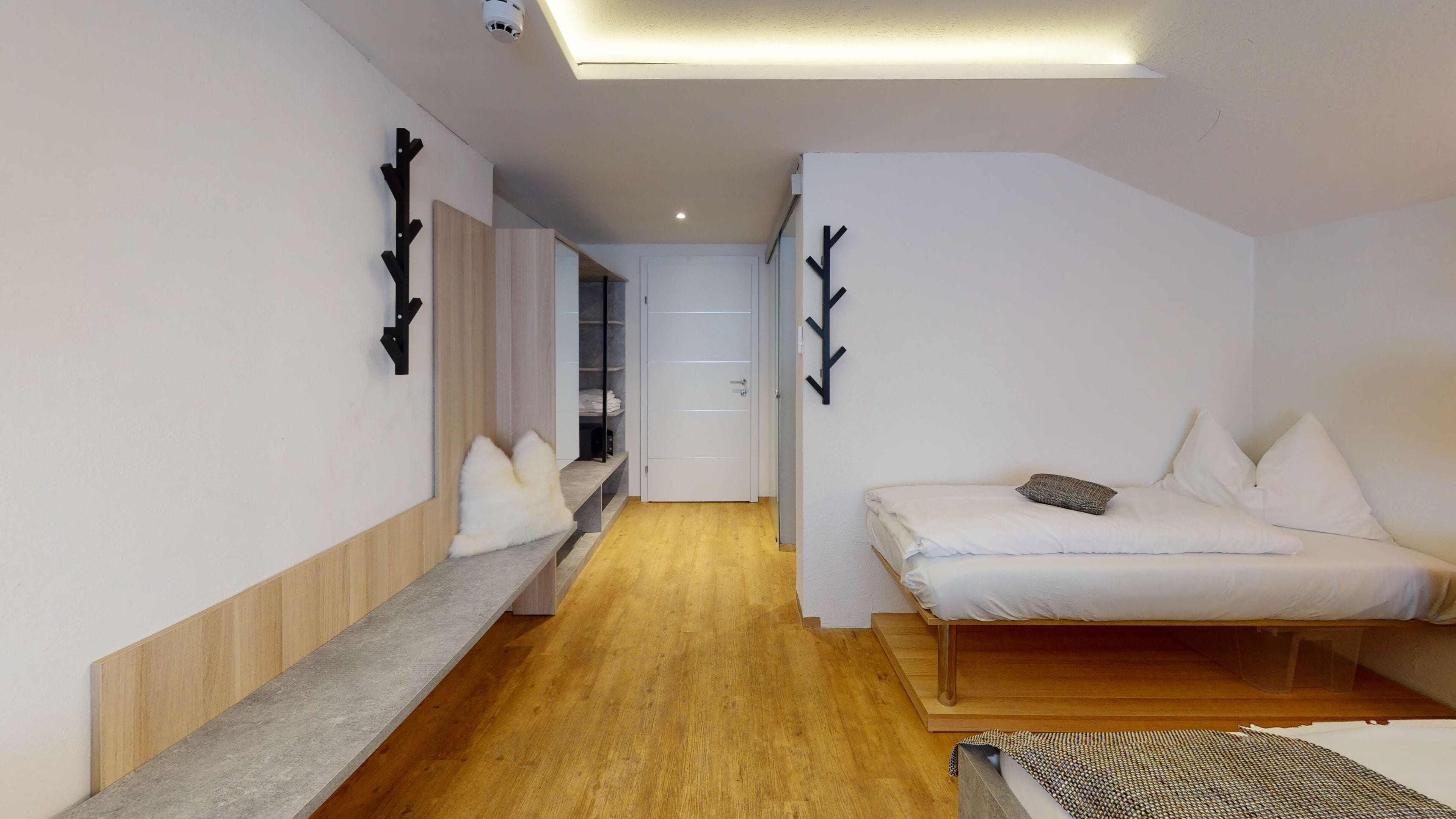 A modern, minimalist hotel room with a wooden floor, a bed with white bedding, a small seating area, and a hallway leading to a bathroom.