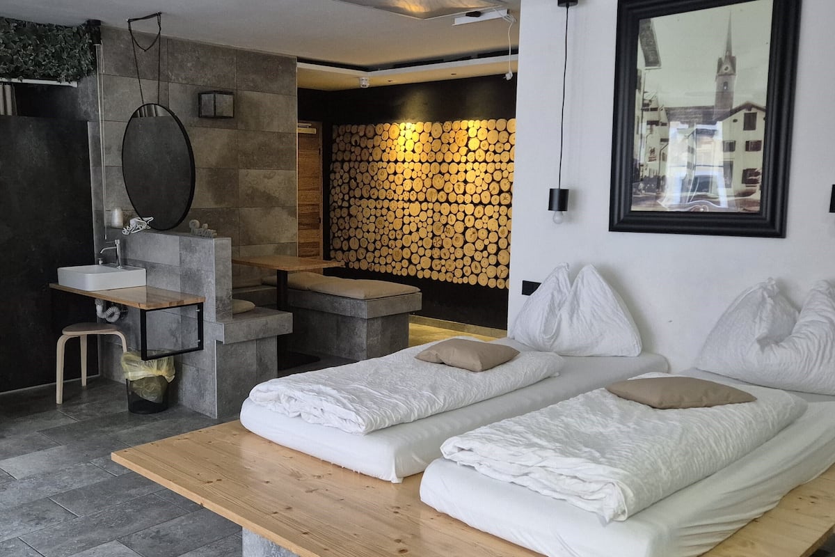 A modern hotel room with a wooden bench, a small sink, a mirror, a bed with white linens, and a wall of stacked logs.