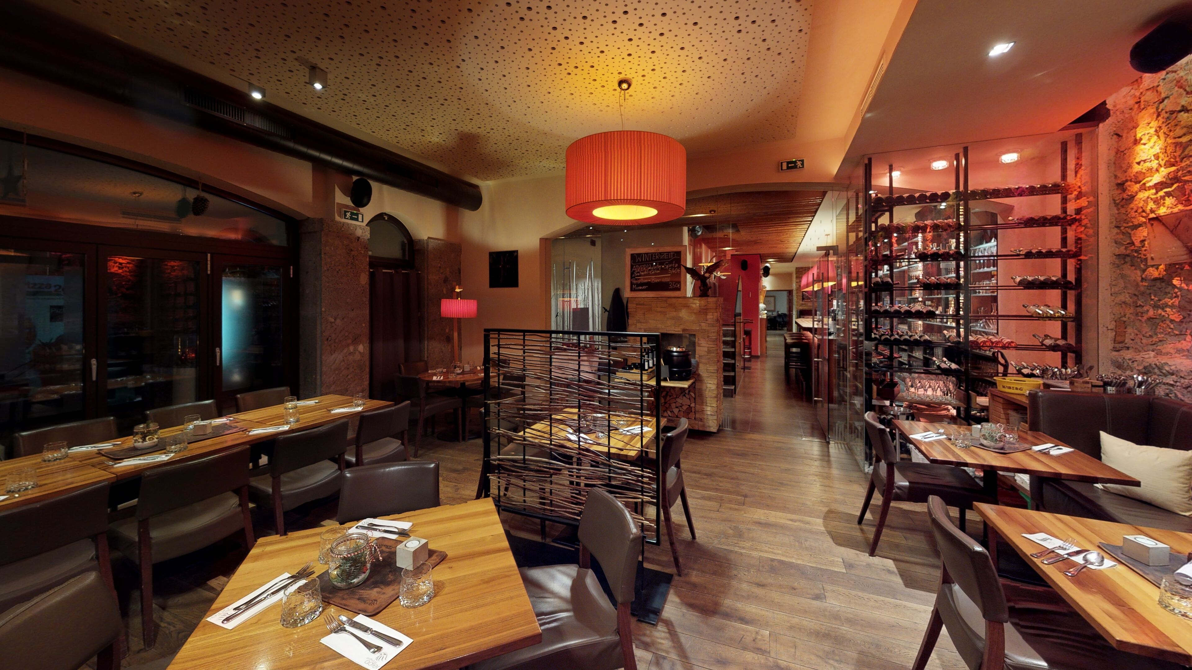 A restaurant interior with wooden tables, chairs, a wine rack, and a red pendant light.