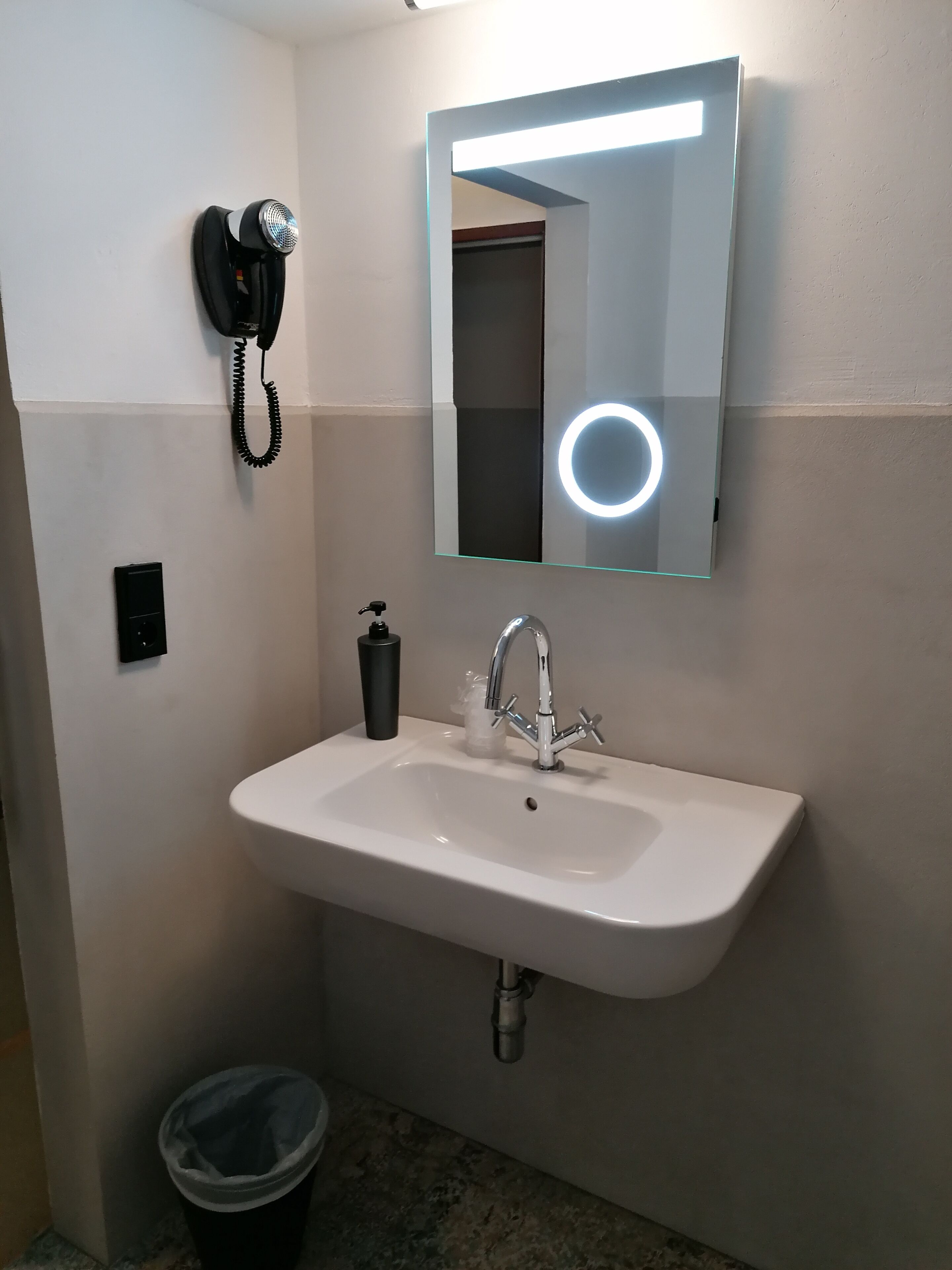 Un baño con lavamanos blanco, un espejo con luz, un secador de pelo y un cubo de basura.