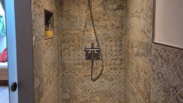 Une douche équipée d’un pommeau de douche à main et d’un robinet mural, installée dans une pièce dont les murs sont recouverts de carreaux à motifs et où l’on peut voir la porte.