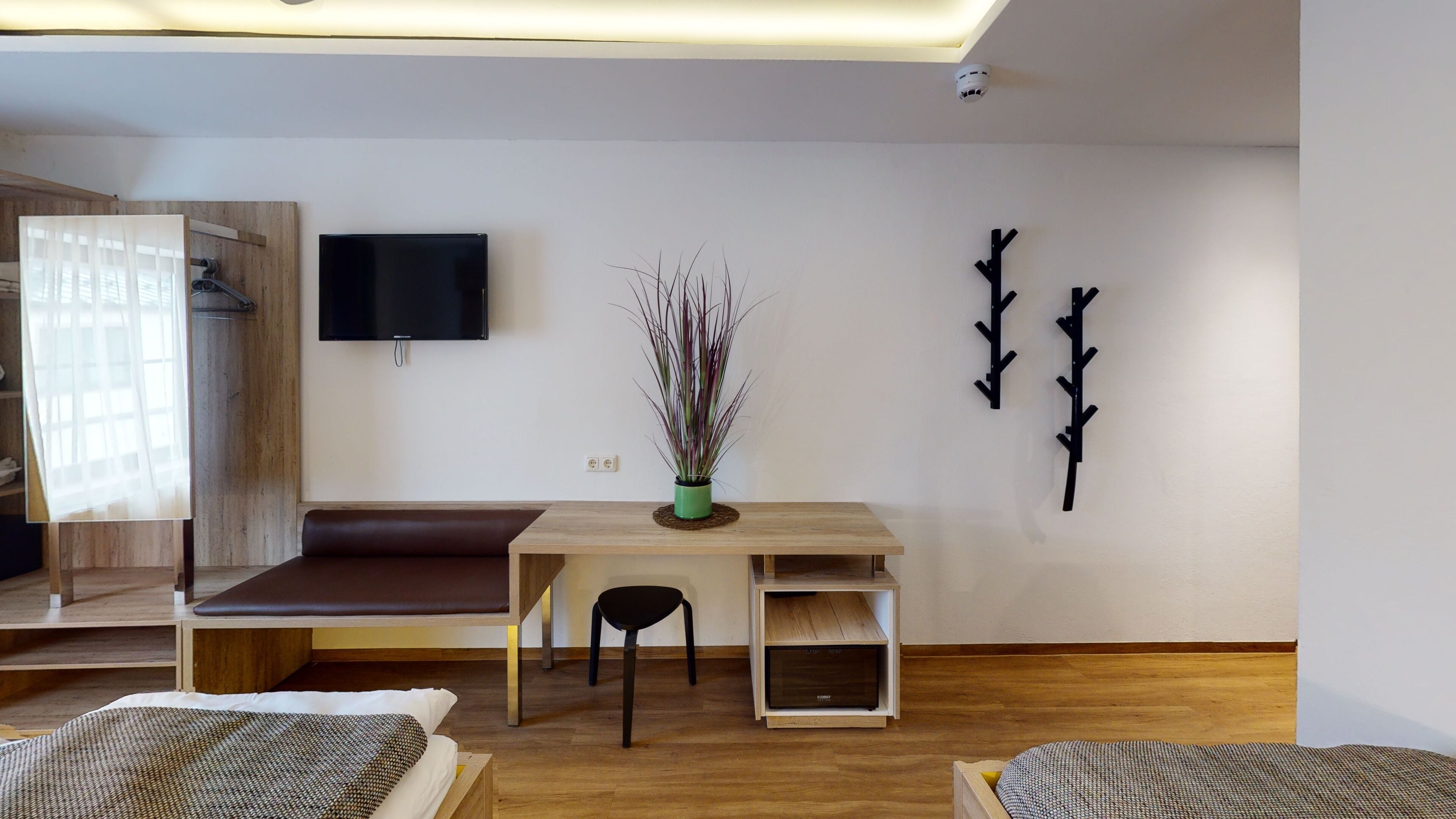 A modern living room with a wooden bench, a small table with a chair, a flat-screen TV mounted on the wall, and a decorative plant in a green pot.