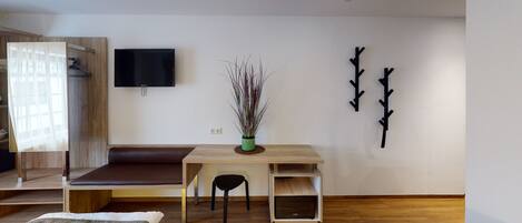 A modern living room with a wooden bench, a small table with a chair, a flat-screen TV mounted on the wall, and a decorative plant in a green pot.