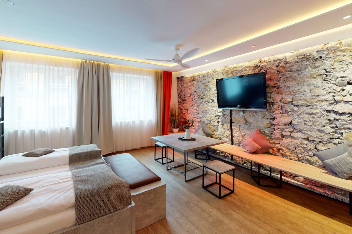 A modern hotel room with a bed, a desk, a bench, a TV, and a stone wall.