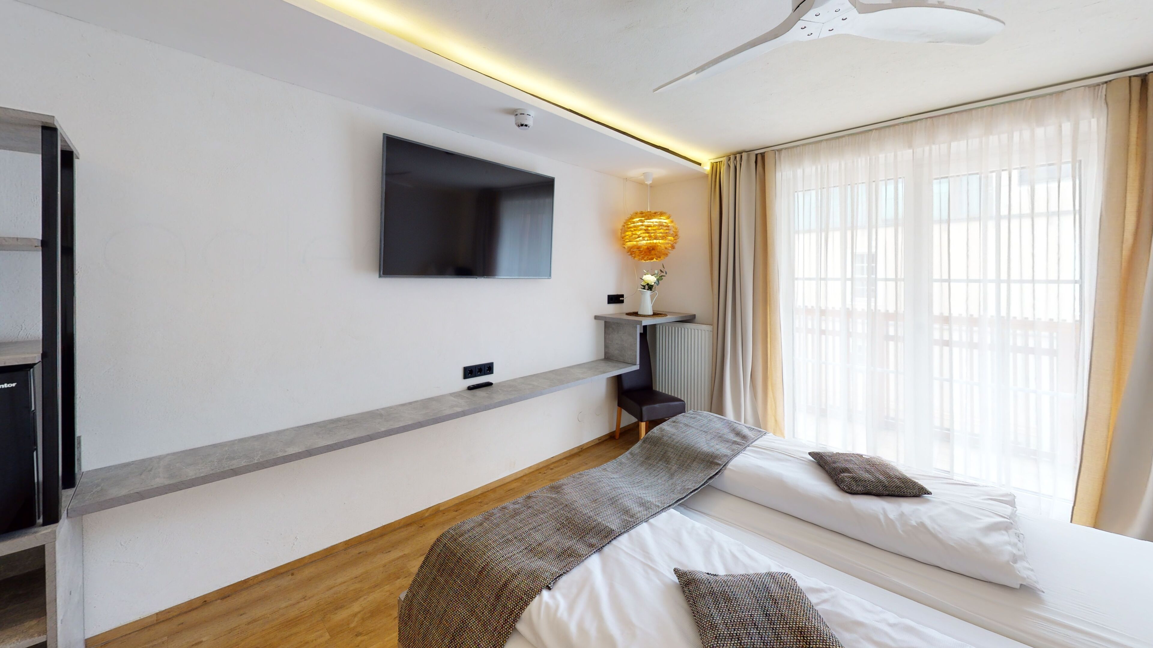 A modern hotel room with a large bed, a flat-screen TV mounted on the wall, a bedside table with a lamp, and a window with sheer curtains.