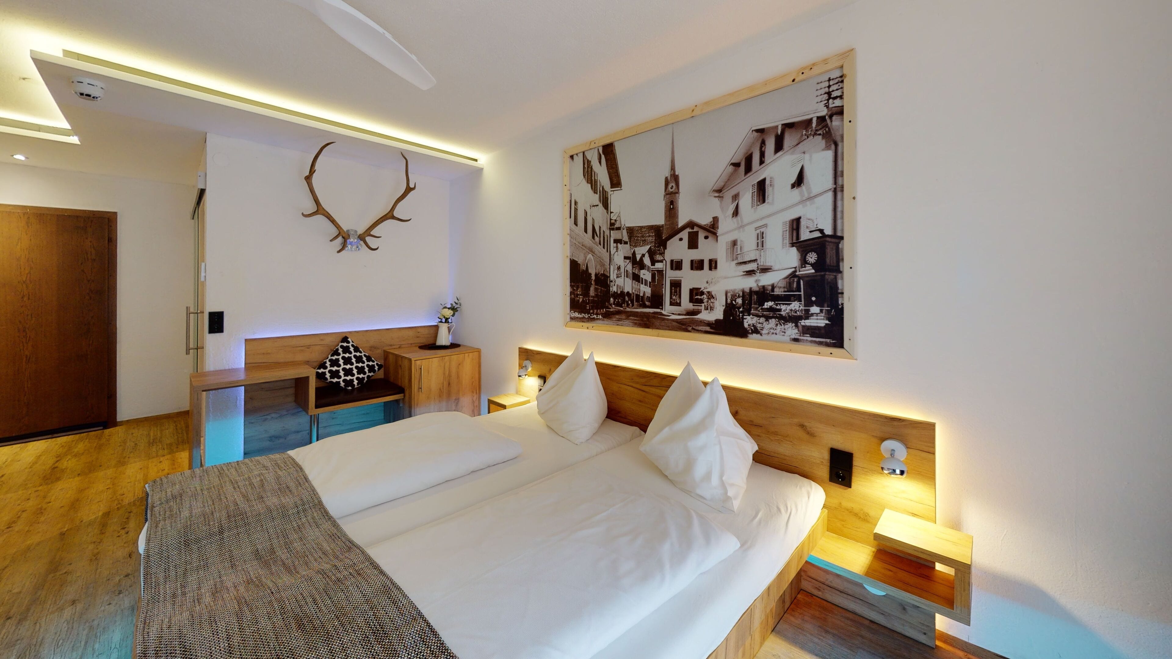 Una habitación de hotel moderna con una cama grande, mesitas de noche, un cabecero de madera, una cornamenta de venado montada en la pared y un cuadro de un paisaje urbano en blanco y negro.