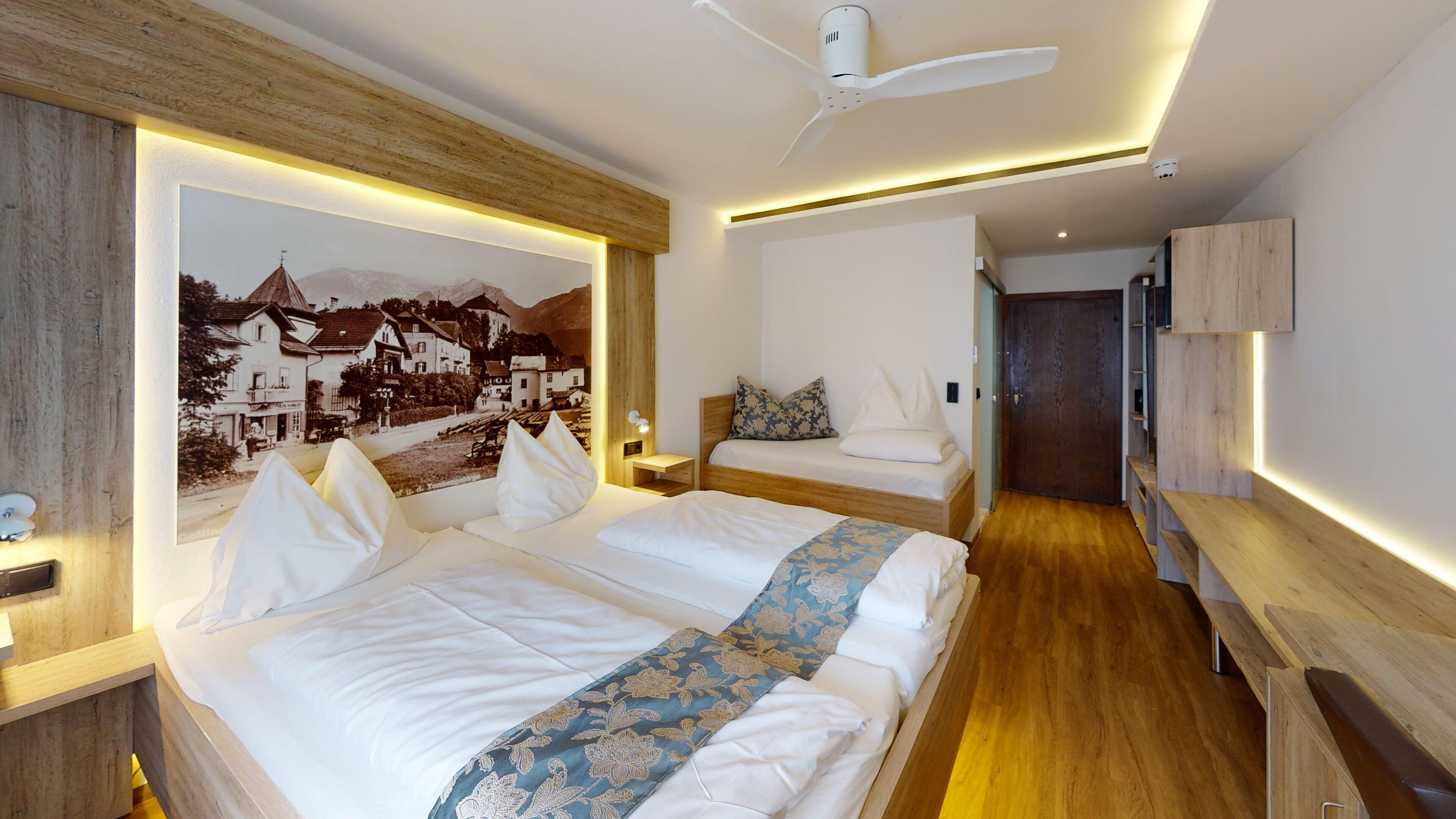 A modern hotel room with a large bed, a smaller bed, a desk, and a wooden headboard.