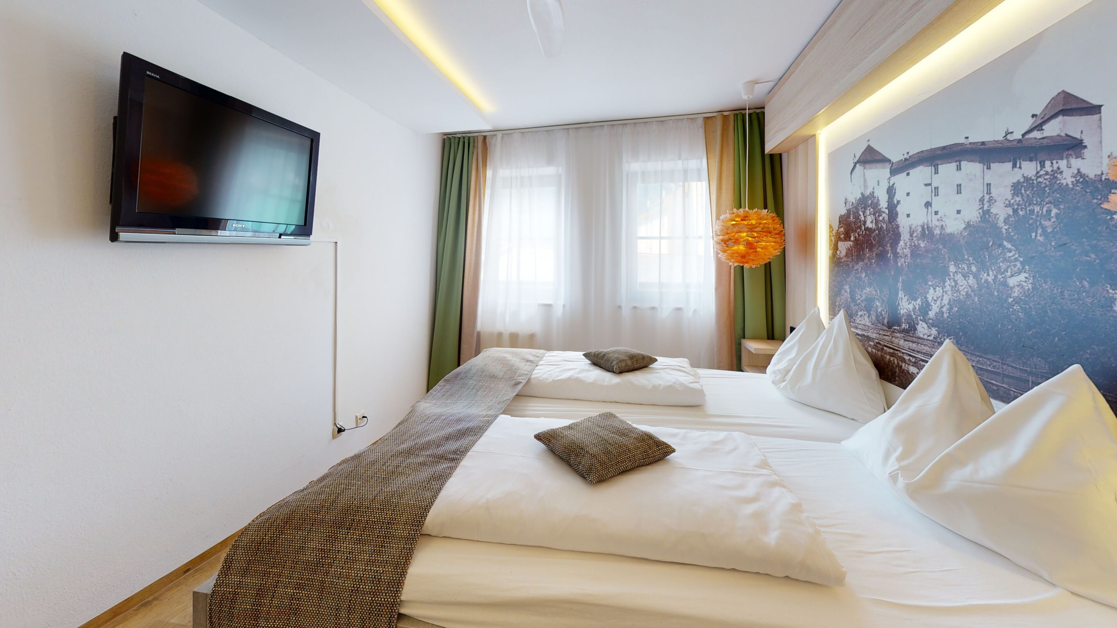 Camera d'albergo con un letto, una TV a parete, un quadro e una finestra con tende.