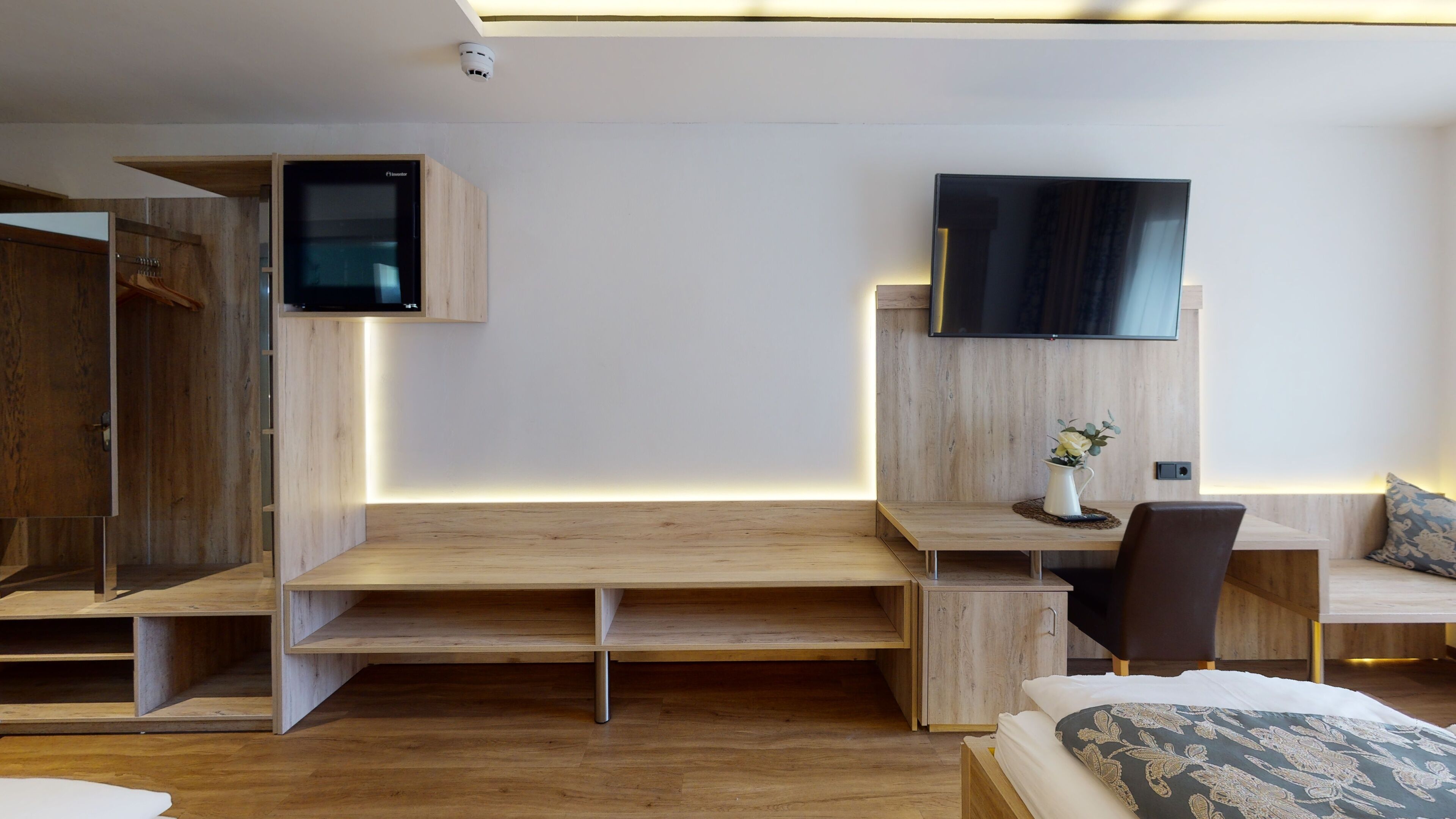 Una habitación de hotel moderna con un escritorio de madera, un televisor de pantalla plana, una cama con ropa de cama estampada y una mesita con un jarrón de flores.
