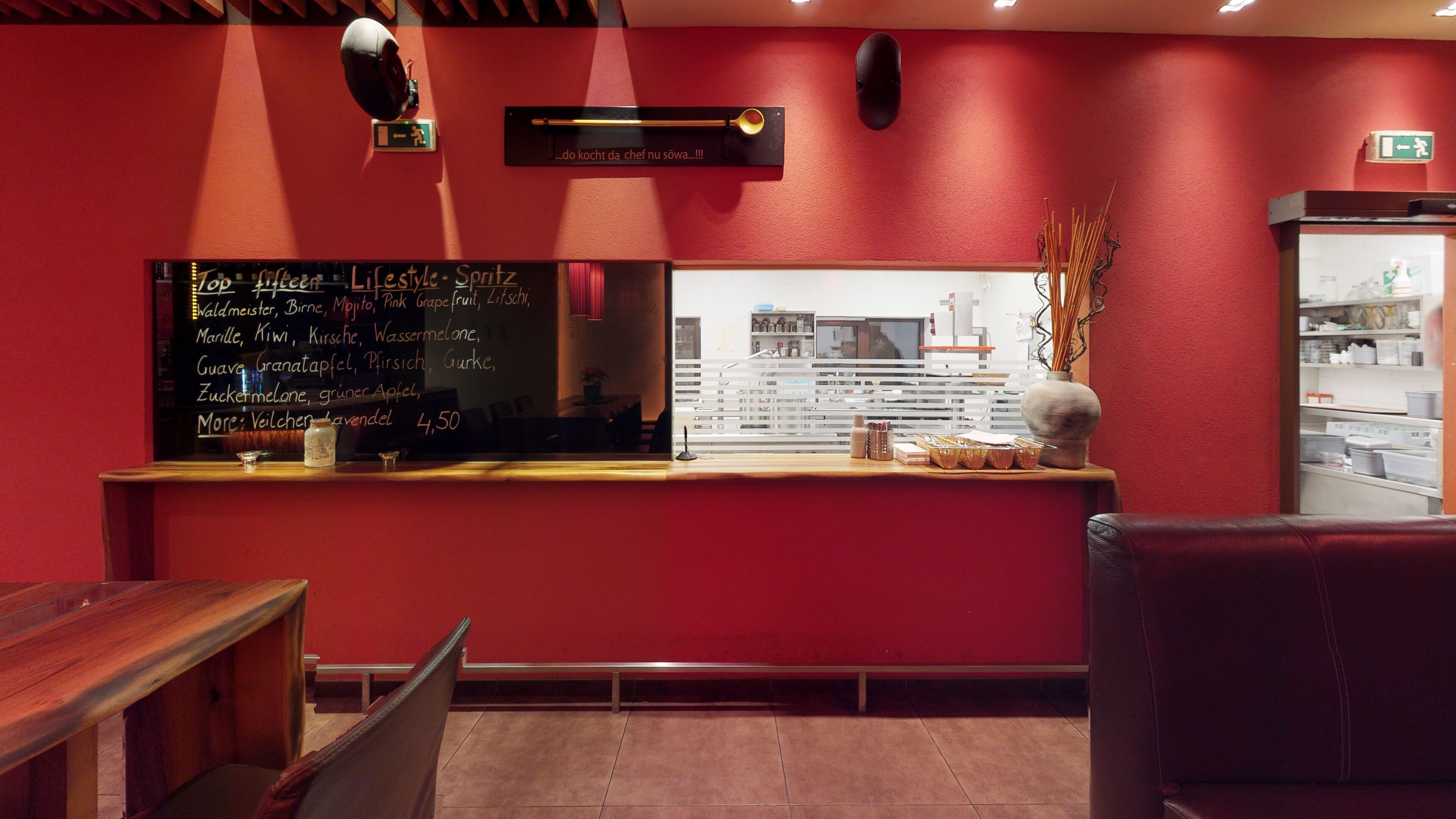 A restaurant interior with a red wall, a chalkboard menu listing various dishes and prices, a wooden counter, and a seating area with a leather chair.