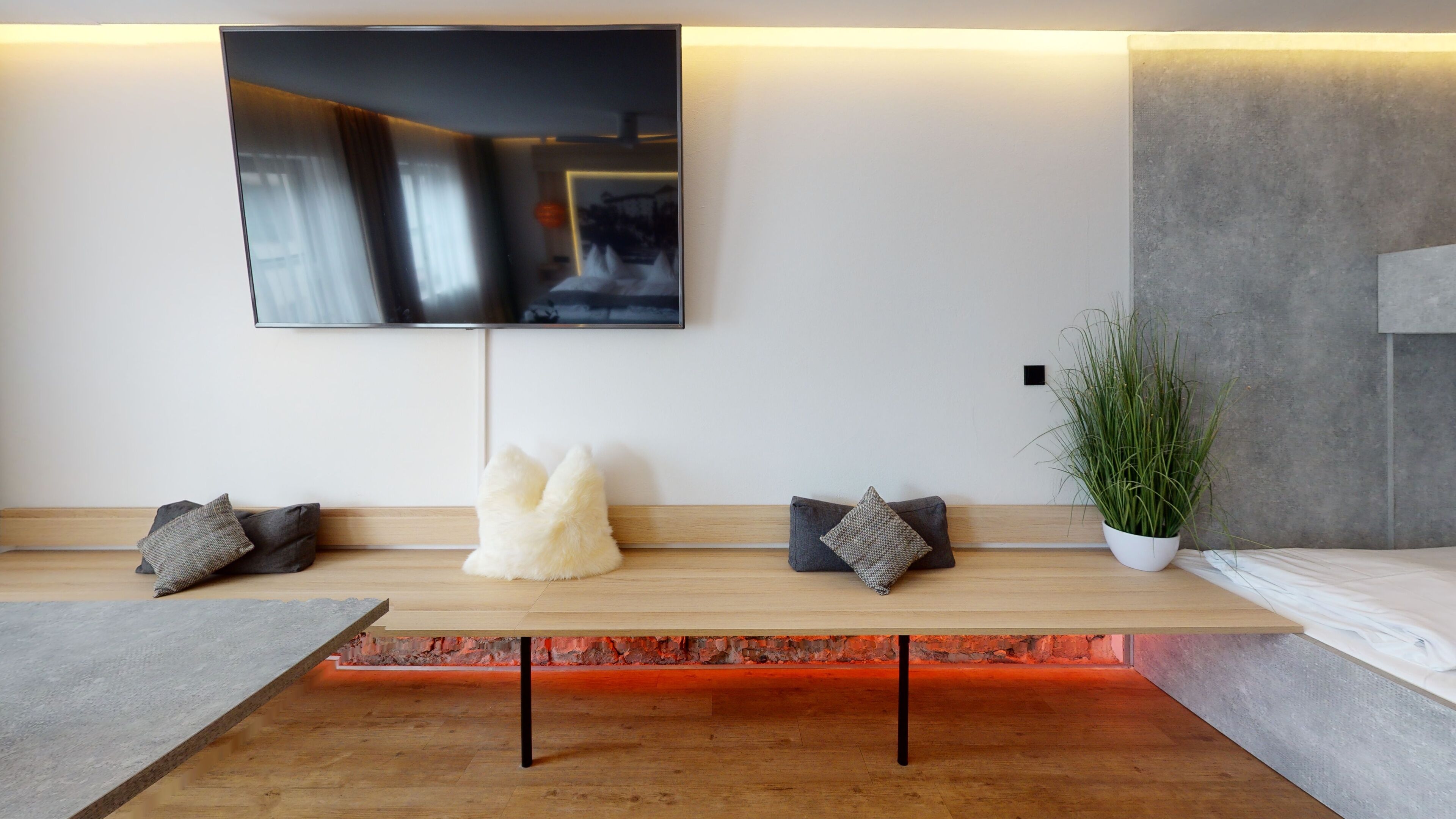 Una sala de estar moderna con un televisor de pantalla plana grande montado en la pared, un banco de madera con cojines, una planta en maceta y una pared de acento de piedra.